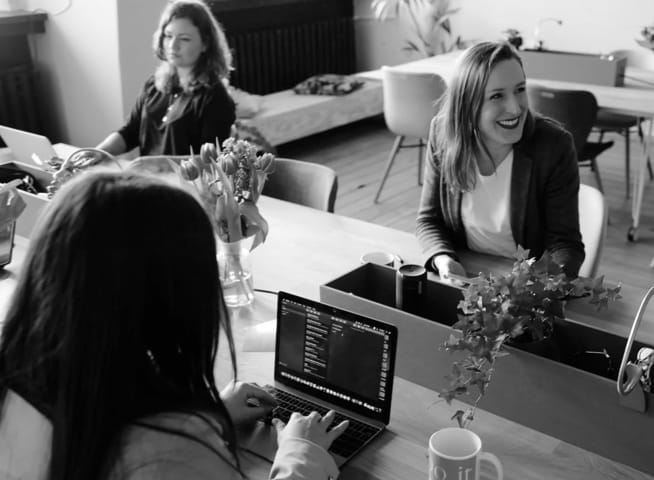 A greyscale photo with purple overlay. Three women in a casual work setting. They are sitting at a table, two are working on laptops and one is smiling to someone off camera.